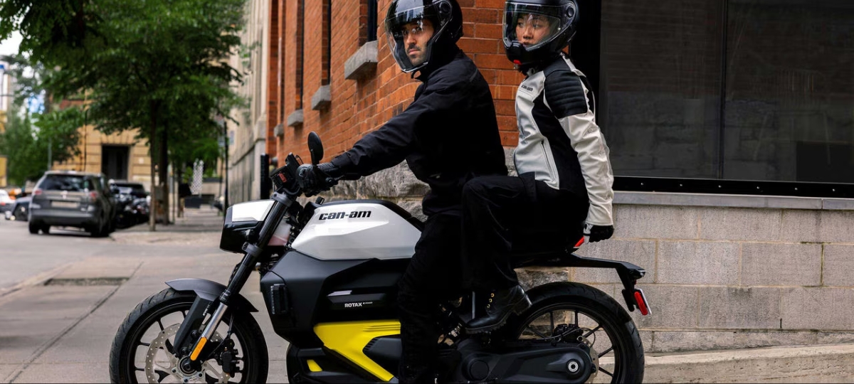 Two riders on a sleek Can-Am EV motorcycle stop in a vibrant urban setting, ready for an eco-friendl…